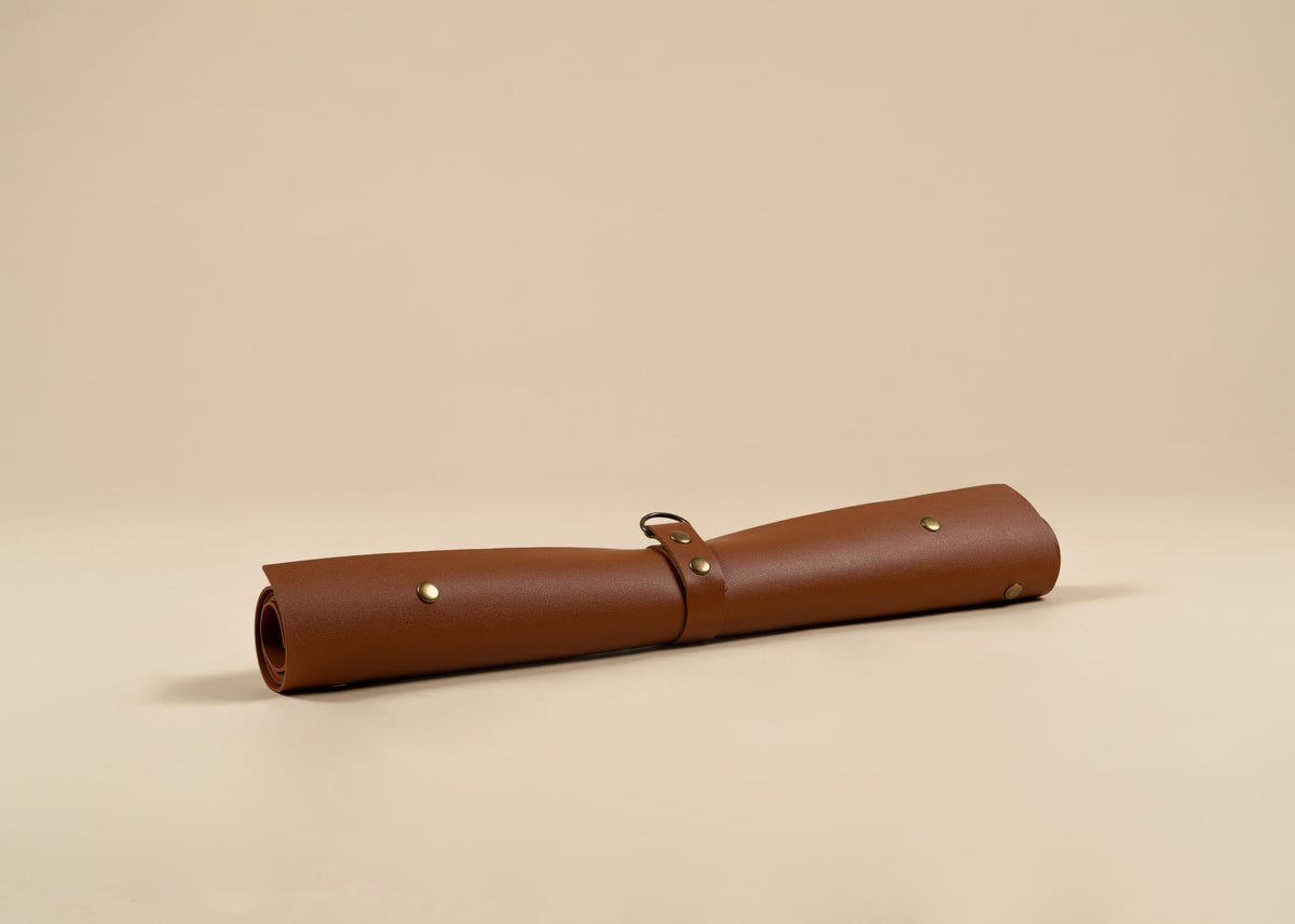 50x75cm in size, A rolled brown leather sleeve or case with brass-tone rivets and a small metal loop closure, photographed on a plain warm beige studio background.