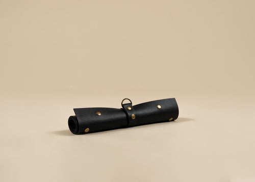 A black, 30x60cm in size,  rolled leather pouch with brass studs and a metal ring, displayed on a warm beige background.