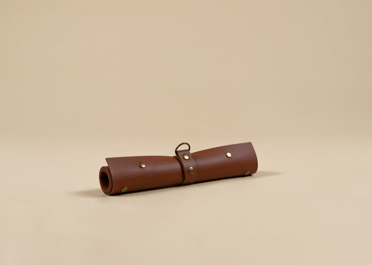 30x60cm in size, A rolled brown leather portfolio or organizer sleeve with brass grommets and a metal loop, resting on a solid beige background.