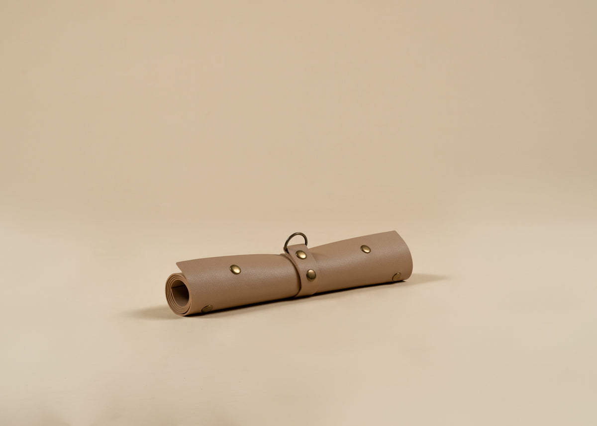30x60cm in size, A rolled-up tan leather pouch or case with brass-tone rivets and a small metal ring, photographed on a plain warm beige studio background.