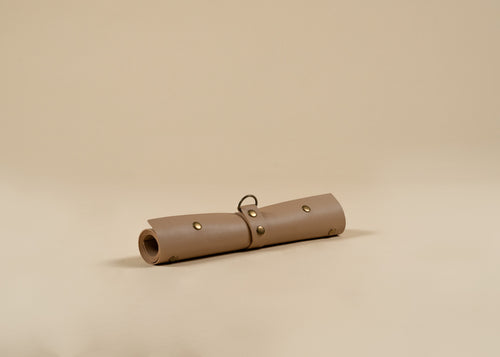 30x60cm in size, A rolled-up tan leather pouch or case with brass-tone rivets and a small metal ring, photographed on a plain warm beige studio background.