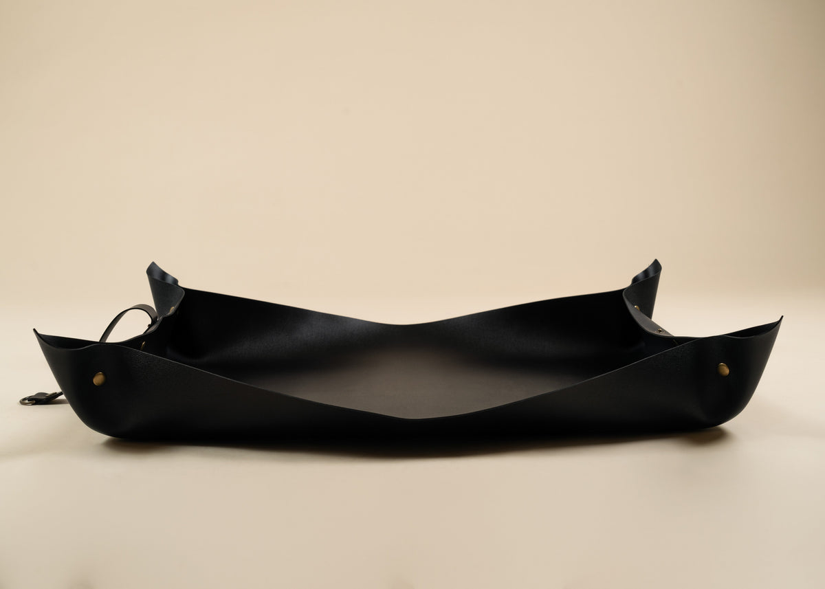 50x75cm in size A black leather boat-shaped tray or bowl with small brass grommets and a hanging loop, photographed on a plain beige background.
