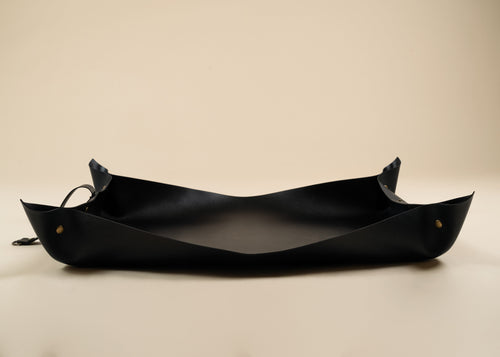 50x75cm in size A black leather boat-shaped tray or bowl with small brass grommets and a hanging loop, photographed on a plain beige background.
