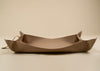 50x75cm in size, A minimalist, boat-shaped leather tray in a warm taupe-brown color, featuring small brass grommets and a matching leather tie on a solid beige background.