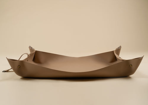 50x75cm in size, A minimalist, boat-shaped leather tray in a warm taupe-brown color, featuring small brass grommets and a matching leather tie on a solid beige background.
