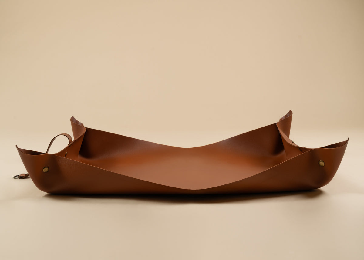 50x75cm in size, A brown leather tray with a curved, boat-like shape and small brass hardware, photographed on a plain beige studio background.