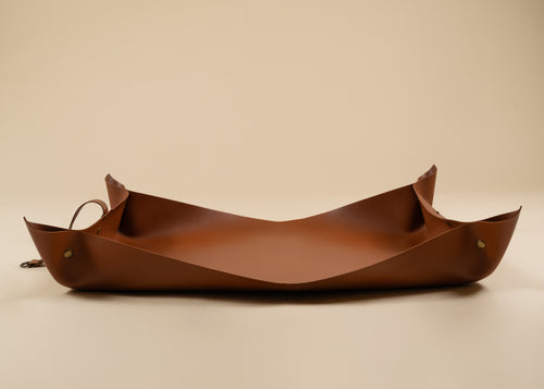 50x75cm in size, A brown leather tray with a curved, boat-like shape and small brass hardware, photographed on a plain beige studio background.