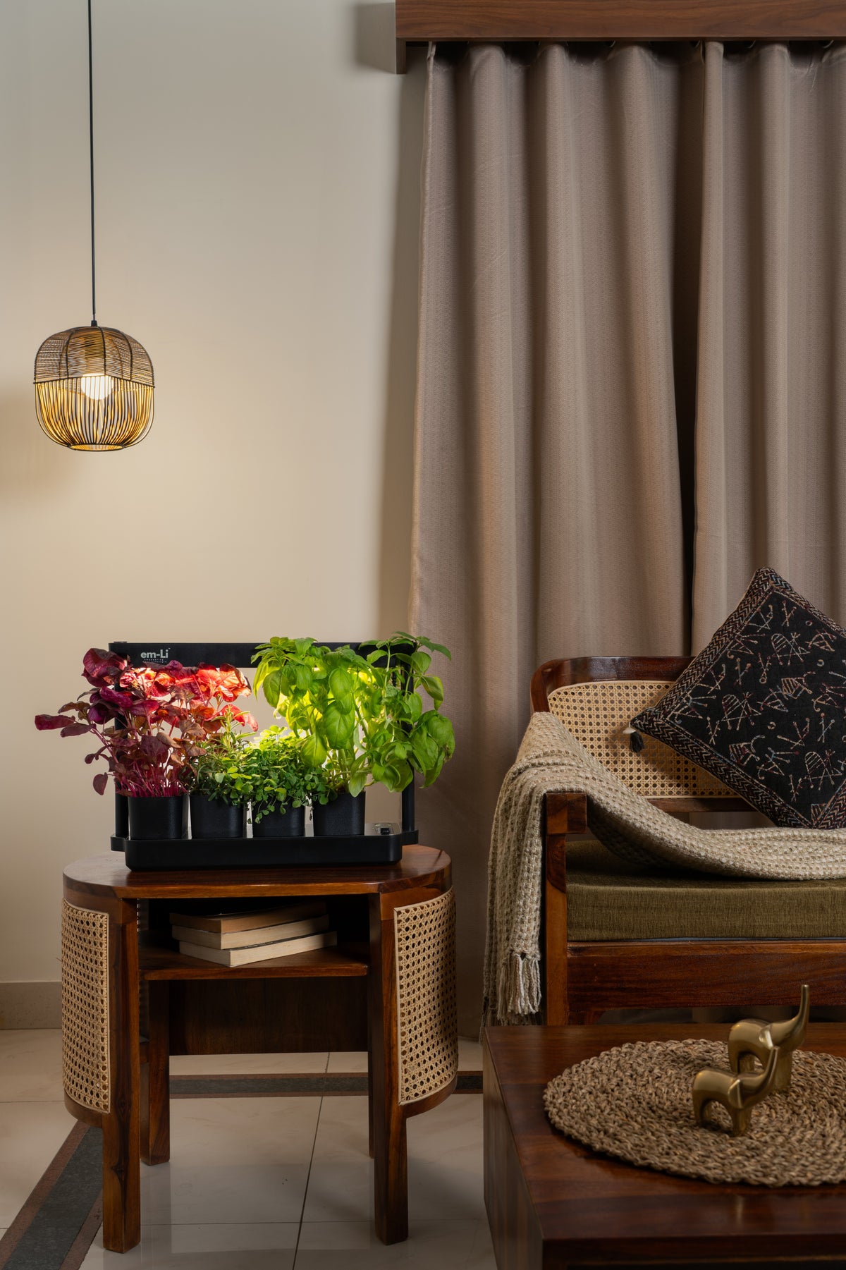 A styled interior scene with a wooden side table holding a black planter box filled with colorful flowers and green leafy plants. A woven pendant light hangs above, and a cushioned wooden armchair with a throw blanket and pillow sits beside tall taupe curtains.