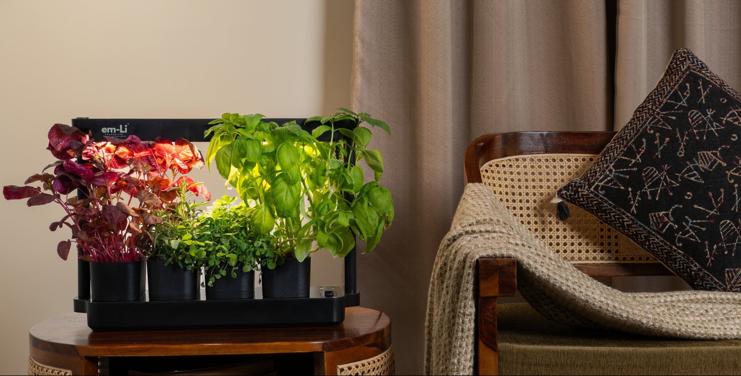 A styled interior scene with a wooden side table holding a black planter box filled with colorful flowers and green leafy plants. A woven pendant light hangs above, and a cushioned wooden armchair with a throw blanket and pillow sits beside tall taupe curtains.