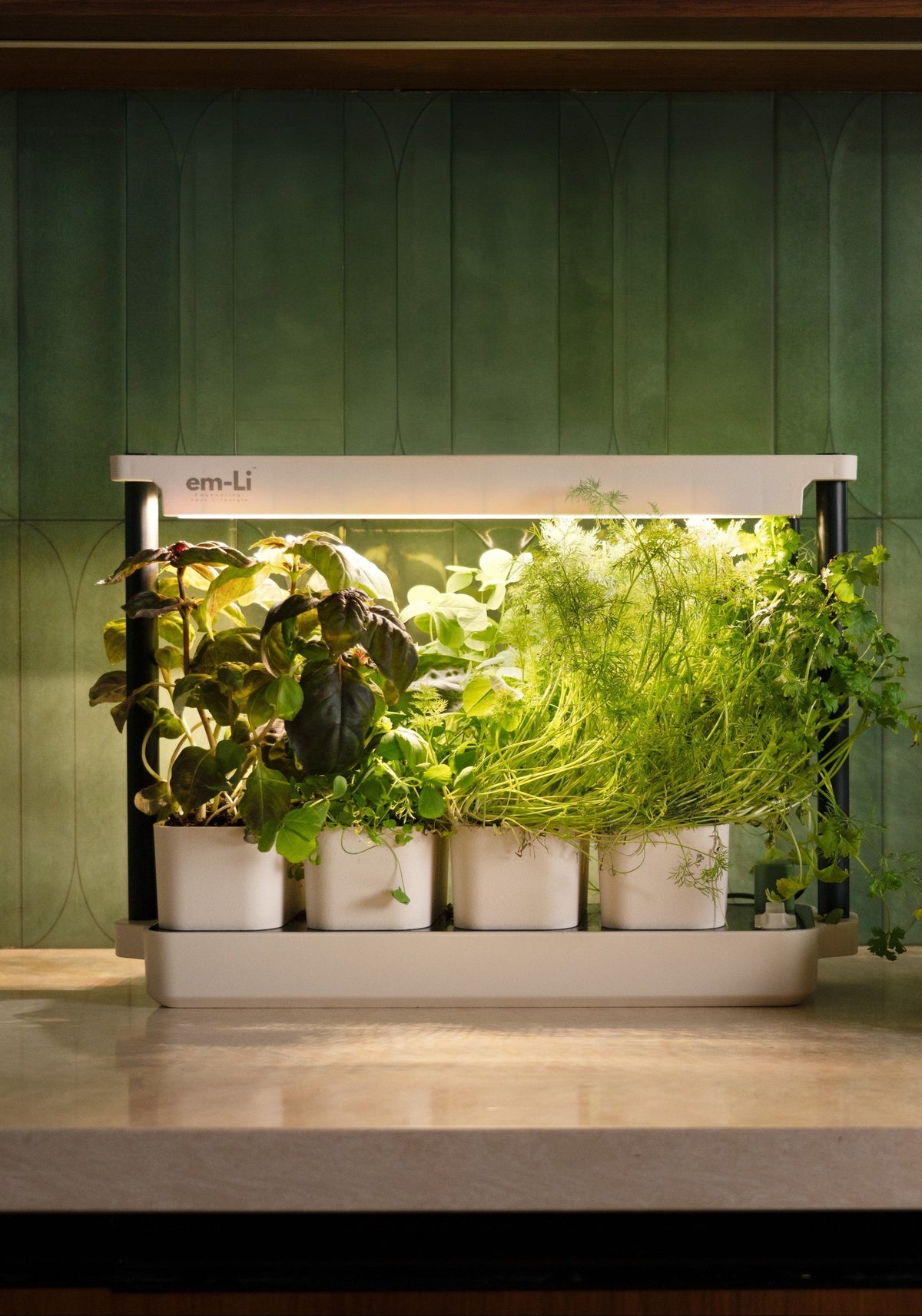 An em-li smart indoor vertical garden system with five white pots under a white light bar, filled with green leafy plants, set on a kitchen countertop against a green tiled backsplash.