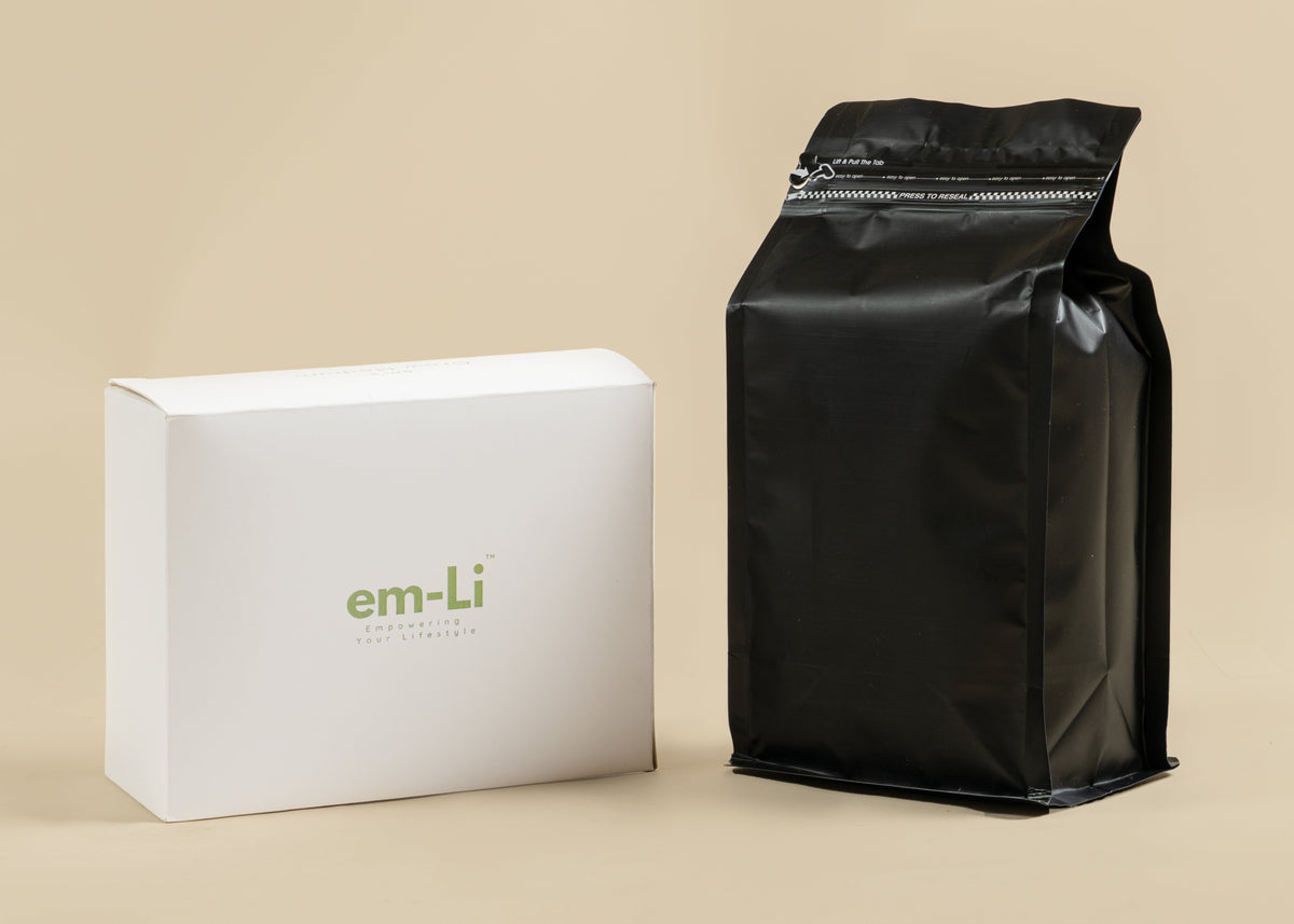 A white rectangular product box with the em-Li logo in green stands next to a glossy black resealable stand-up pouch on a warm beige studio background.