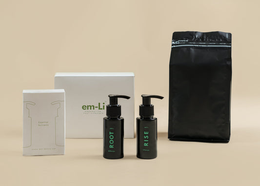 A skincare set on a warm beige background, featuring two black pump bottles labeled ROOT and RISE, a white box with green 'em-Li' branding, a white box with minimalist line art, and a large black resealable pouch.