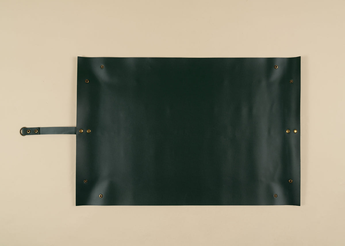 50x75cm in size, A dark forest green leather roll-top canvas or apron with brass grommets and a matching leather strap on the left, laid flat on a beige background.