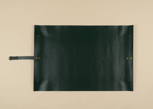 50x75cm in size, A dark forest green leather roll-top canvas or apron with brass grommets and a matching leather strap on the left, laid flat on a beige background.