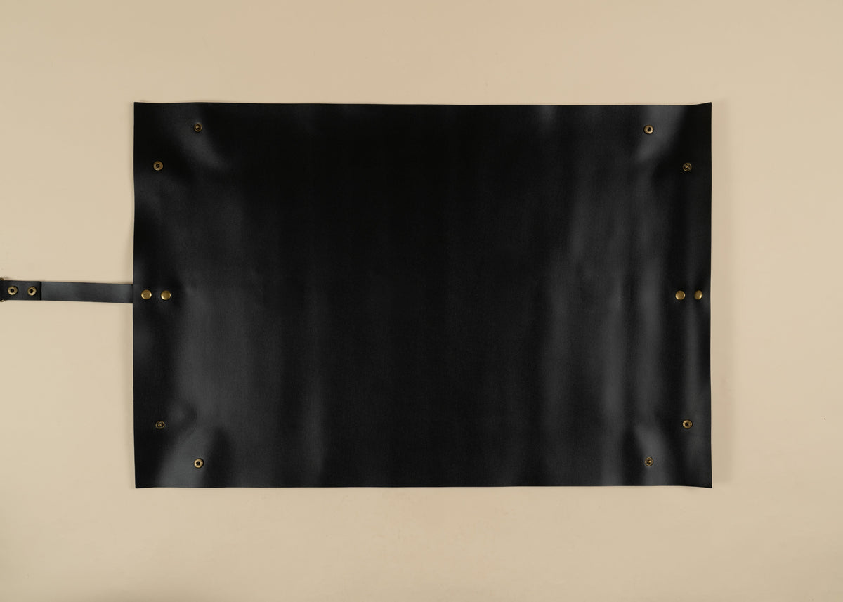 50x75cm in size, A black leather roll-top apron with brass grommets and a leather handle, laid flat against a neutral beige background.