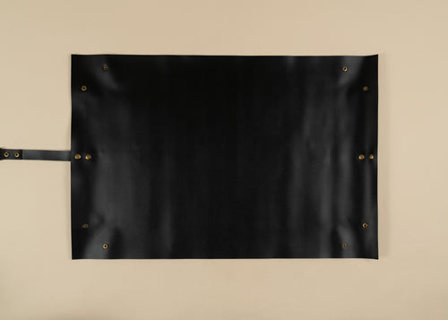 50x75cm in size, A black leather roll-top apron with brass grommets and a leather handle, laid flat against a neutral beige background.