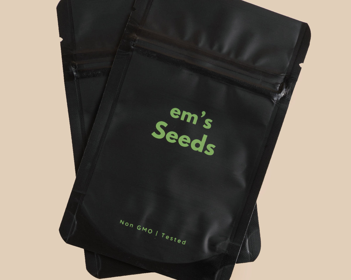 Three black resealable pouches of em's Seeds are stacked on a beige background, with a small square card below reading Coriander Seeds.
