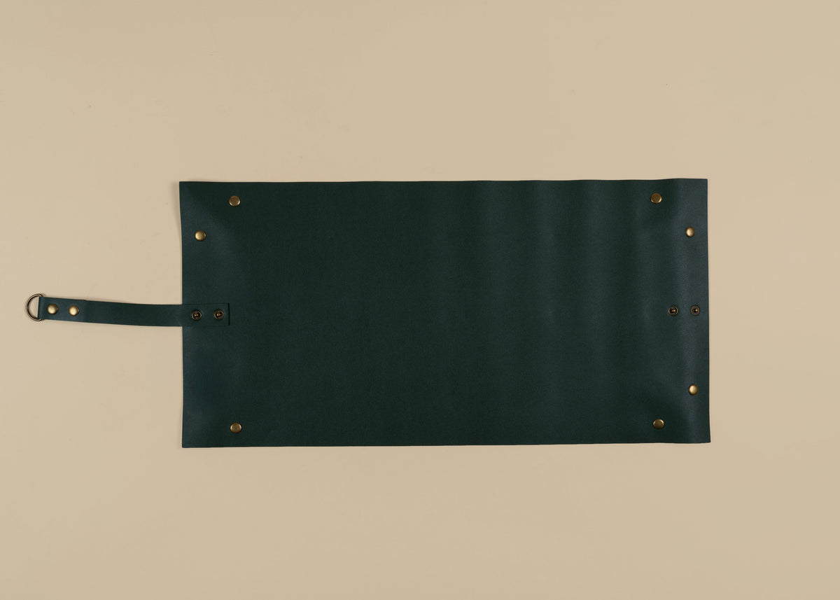 30x60cm in size, A dark forest green leather roll-top tool roll with brass grommets and a matching leather strap on a light beige background