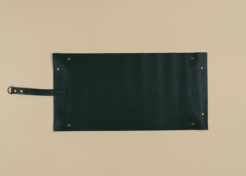 30x60cm in size, A dark forest green leather roll-top tool roll with brass grommets and a matching leather strap on a light beige background