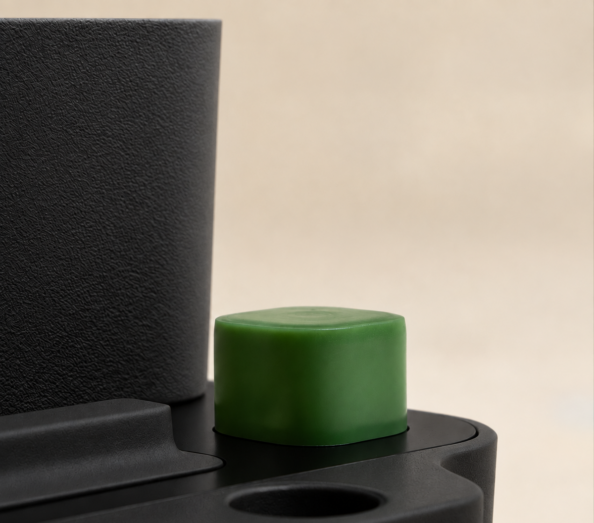 A close-up of a small matte green cylindrical cap or plug resting on a black textured surface, with a blurred warm beige background.