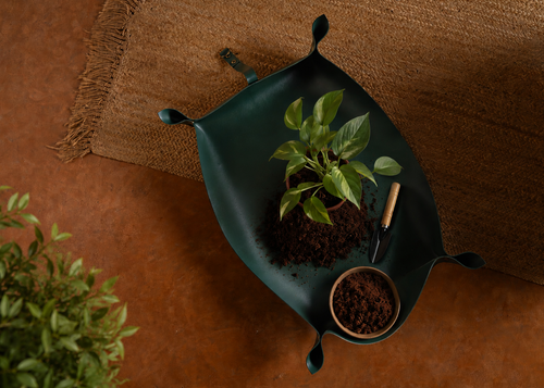A dark green leaf-shaped planter tray holding soil, a small potted plant, and a small trowel, styled on a brown textured rug over a terracotta floor.