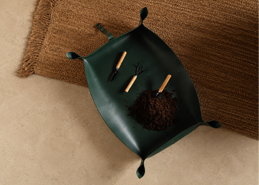 A dark green leaf-shaped planter tray with small hand tools and soil, styled on a beige floor next to a brown woven rug.