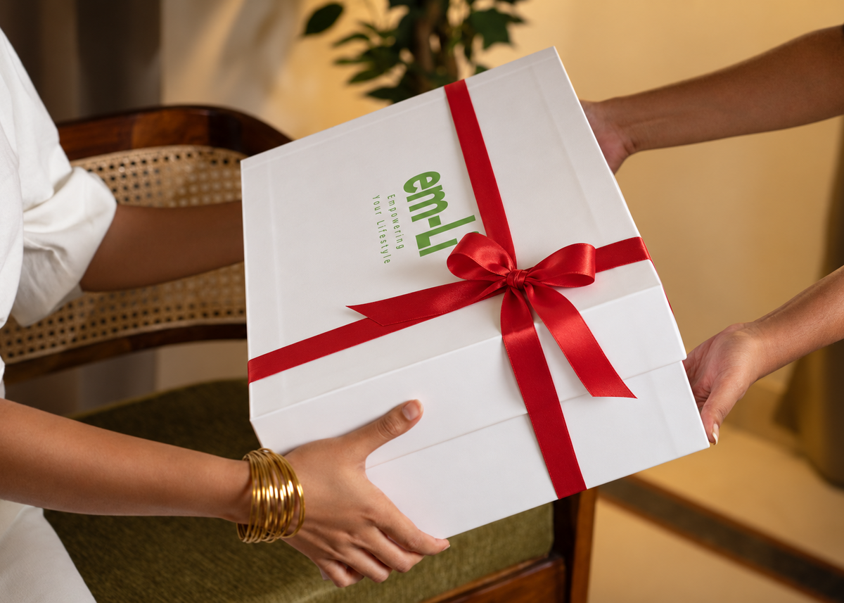 A white rectangular gift box with green 'em-ji' branding, wrapped with a red ribbon and bow, being handed from one person to another in an indoor setting.